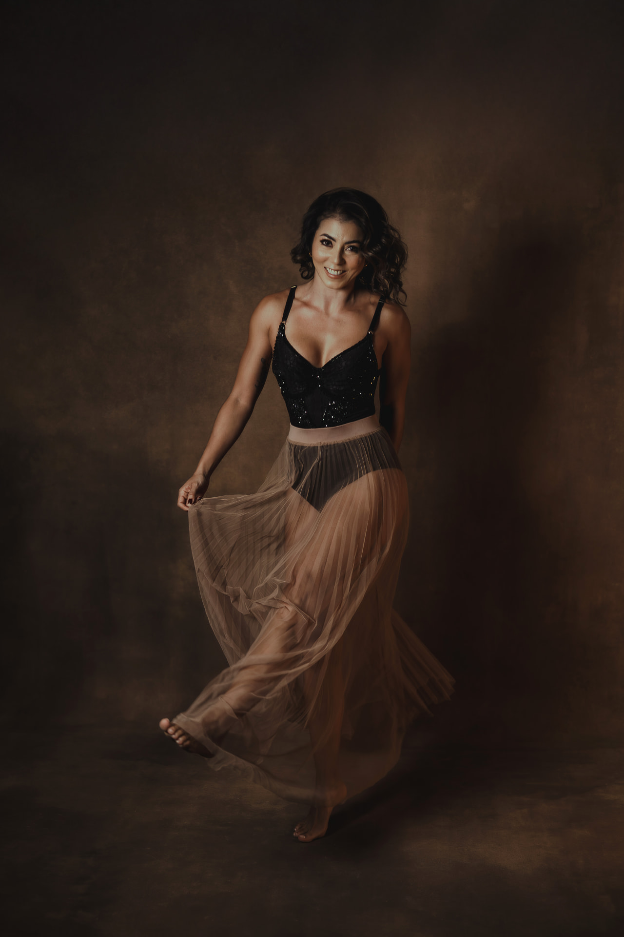 Laura in a black top and sheer pleated skirt against a warm dark backdrop